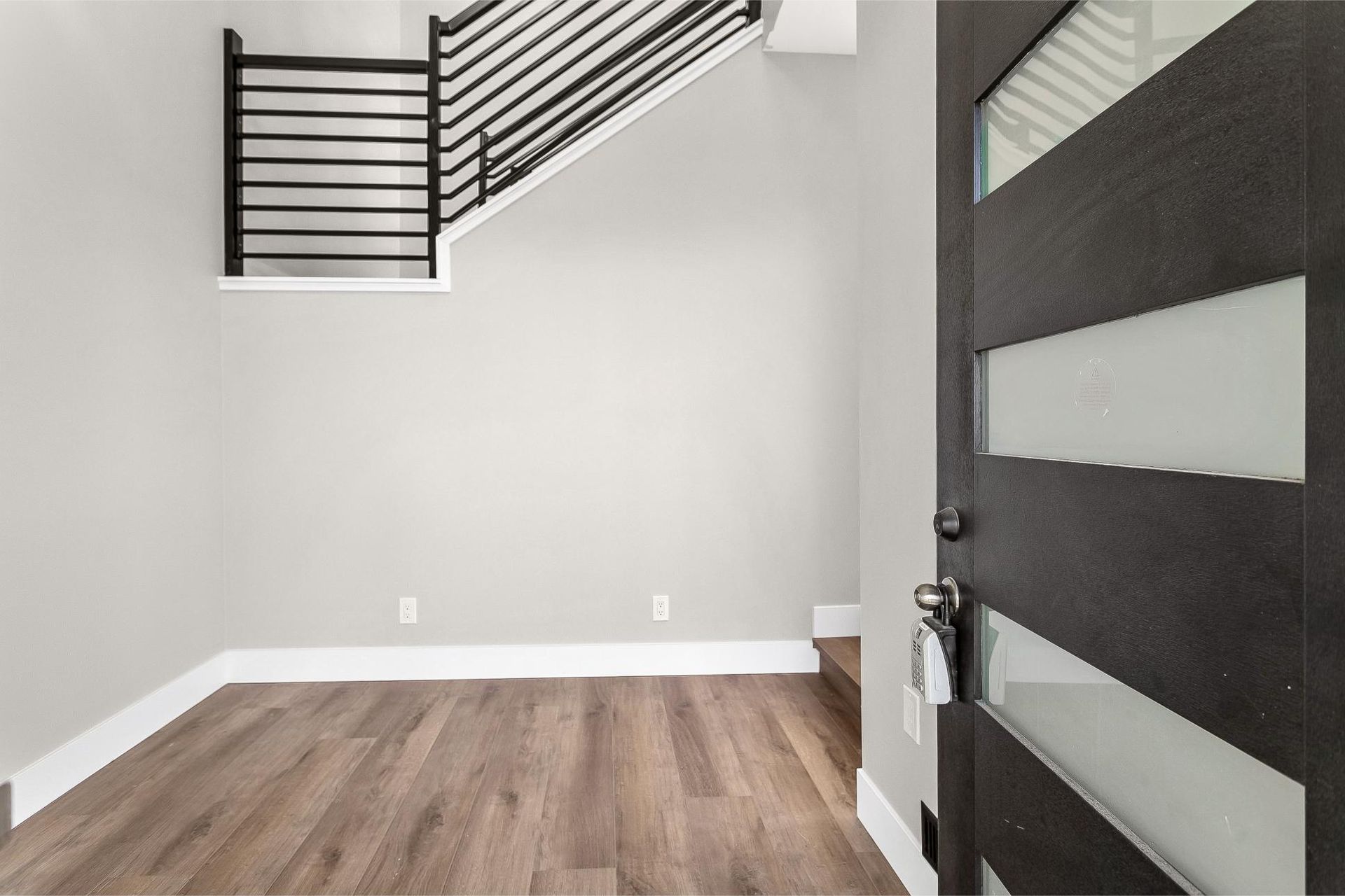 An entryway with light gray walls, wood-look flooring, a modern staircase with black railings, and a dark glass-paneled door.