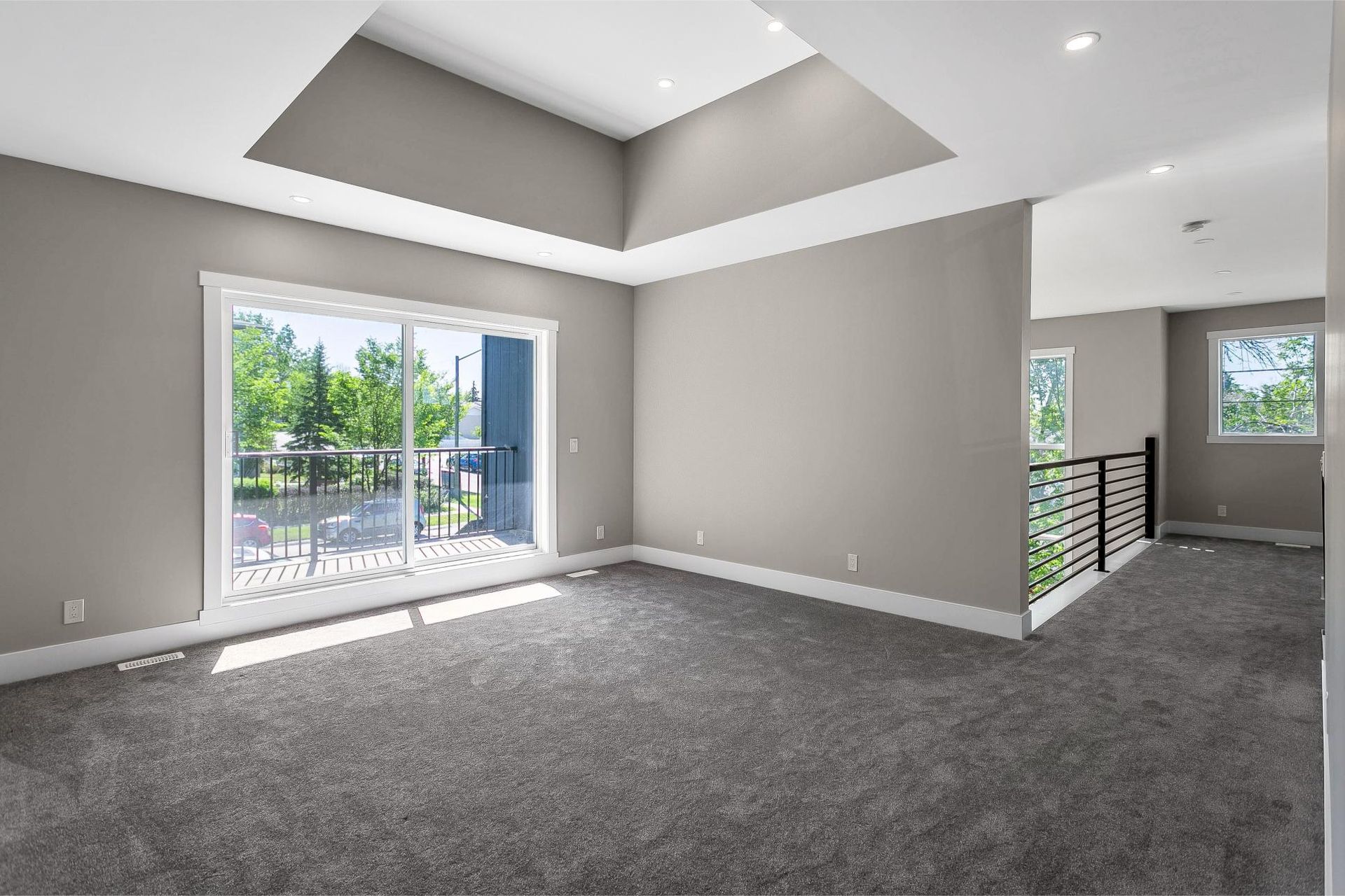 A spacious, empty room with grey carpet, taupe walls, recessed lighting, a large window, and a staircase landing.