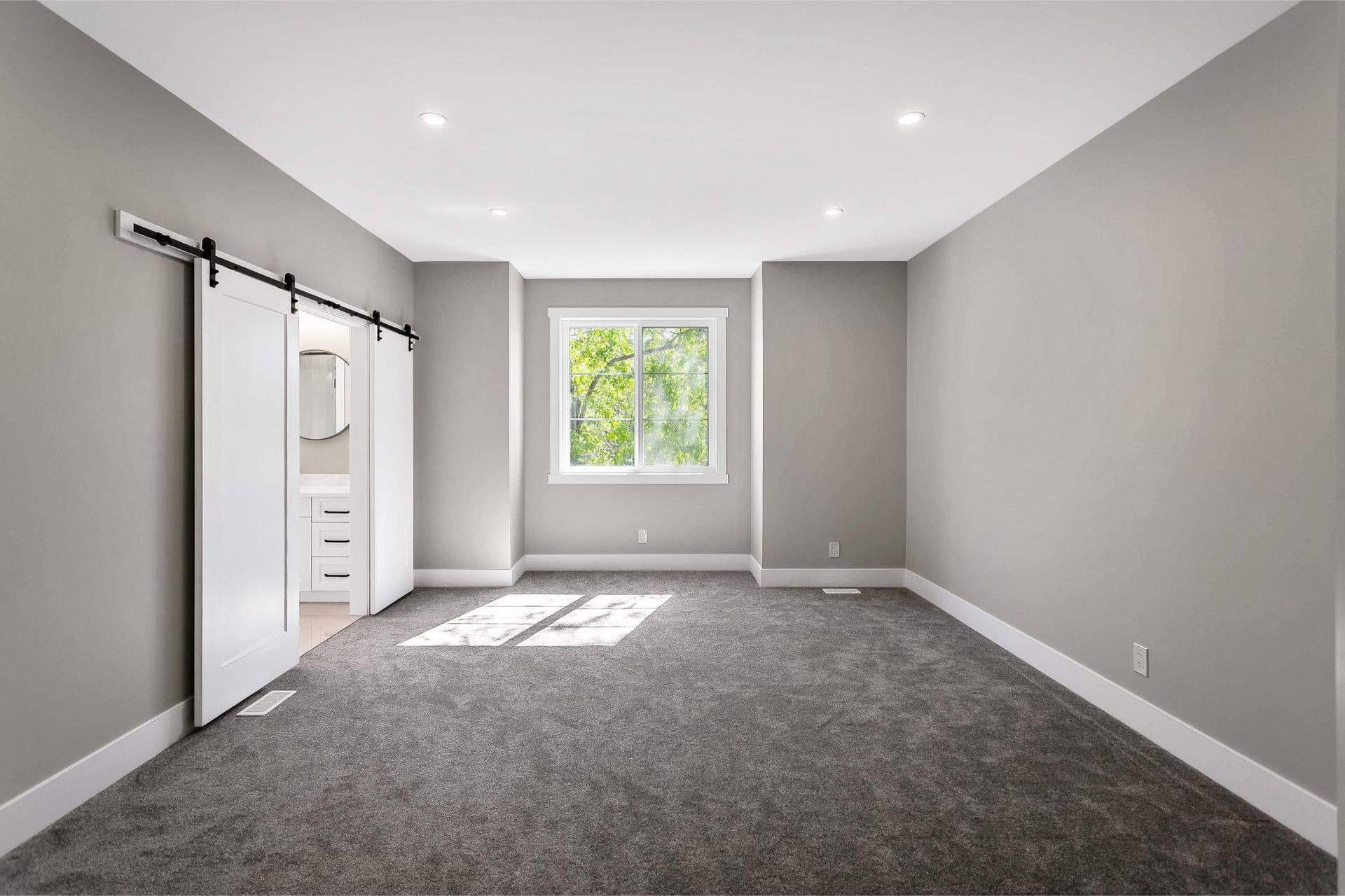 A vacant bedroom with light gray walls, gray carpeting, a single window, and white sliding barn doors leading to a bathroom.