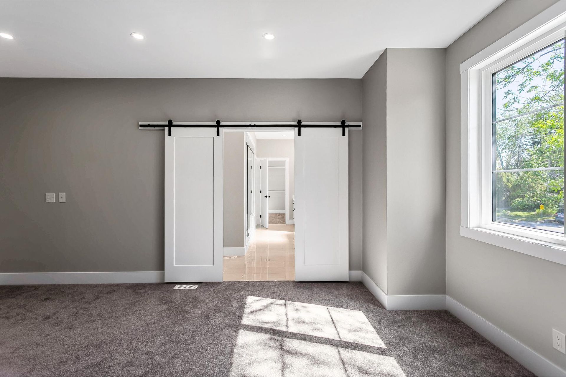 An interior view of a gray-walled room with a double white sliding barn door opening into a hallway and a large window.