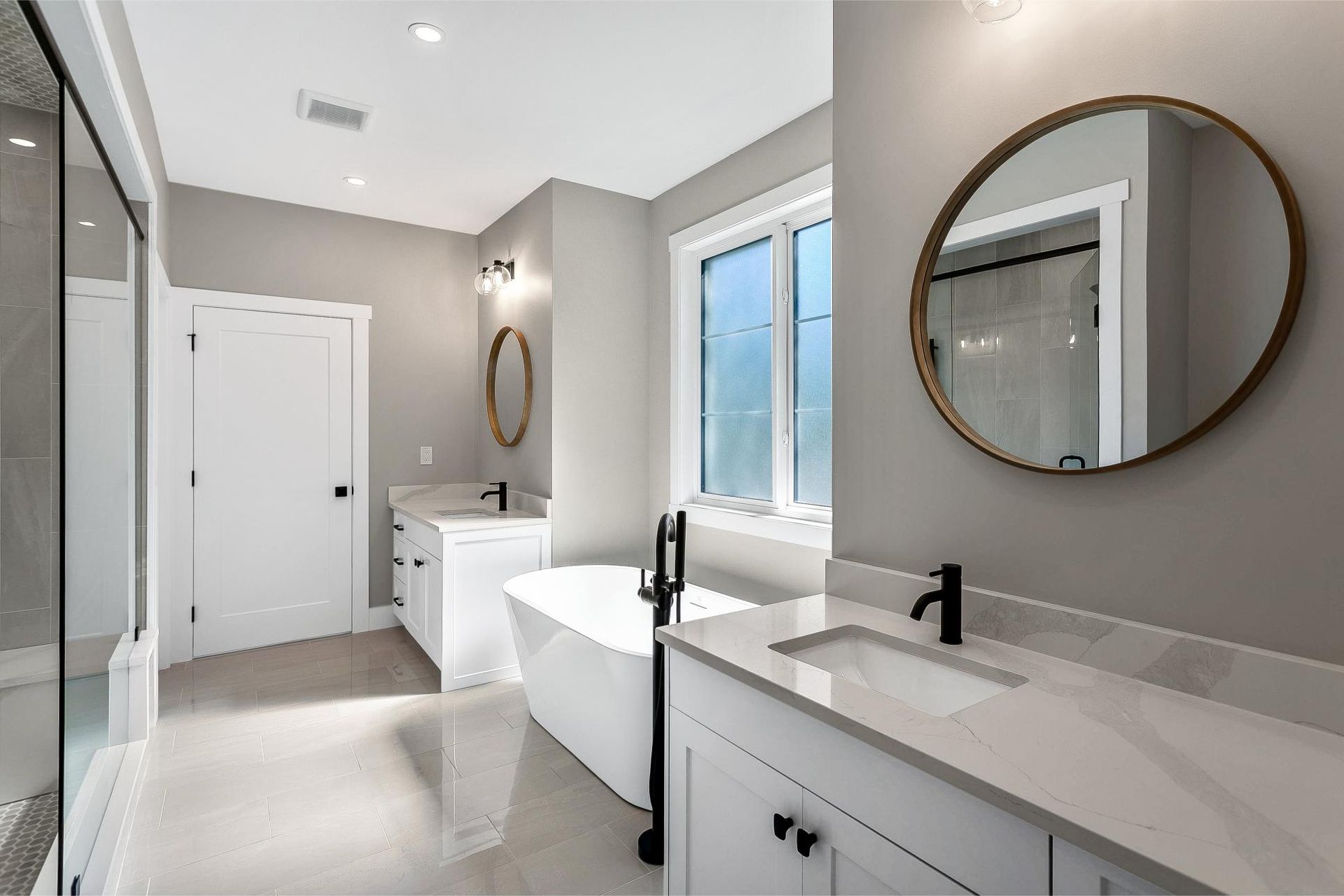 A bright, modern bathroom featuring a white vanity with black fixtures, a freestanding tub, and a round wall mirror.