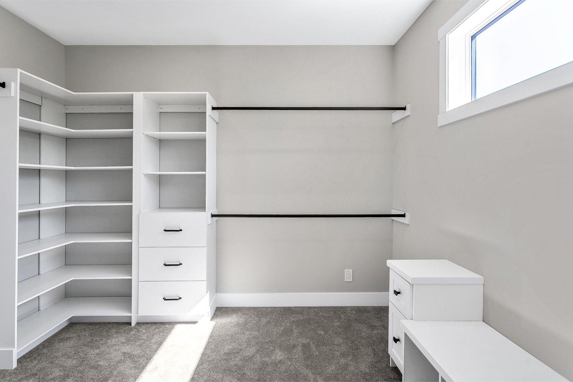 A walk-in closet with white shelving, a chest of drawers, two hanging rods, and a white bench on grey carpet.