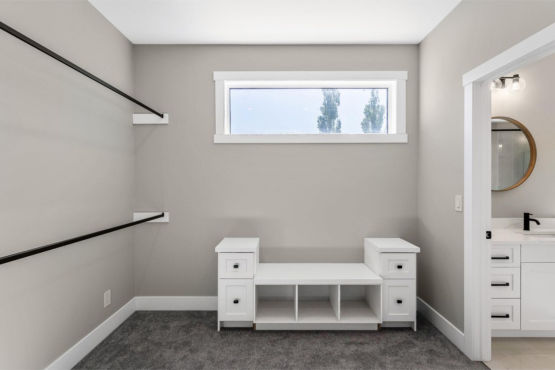 A walk-in closet with gray carpet, two black wall-mounted clothing rods, a white storage bench, and a rectangular window.