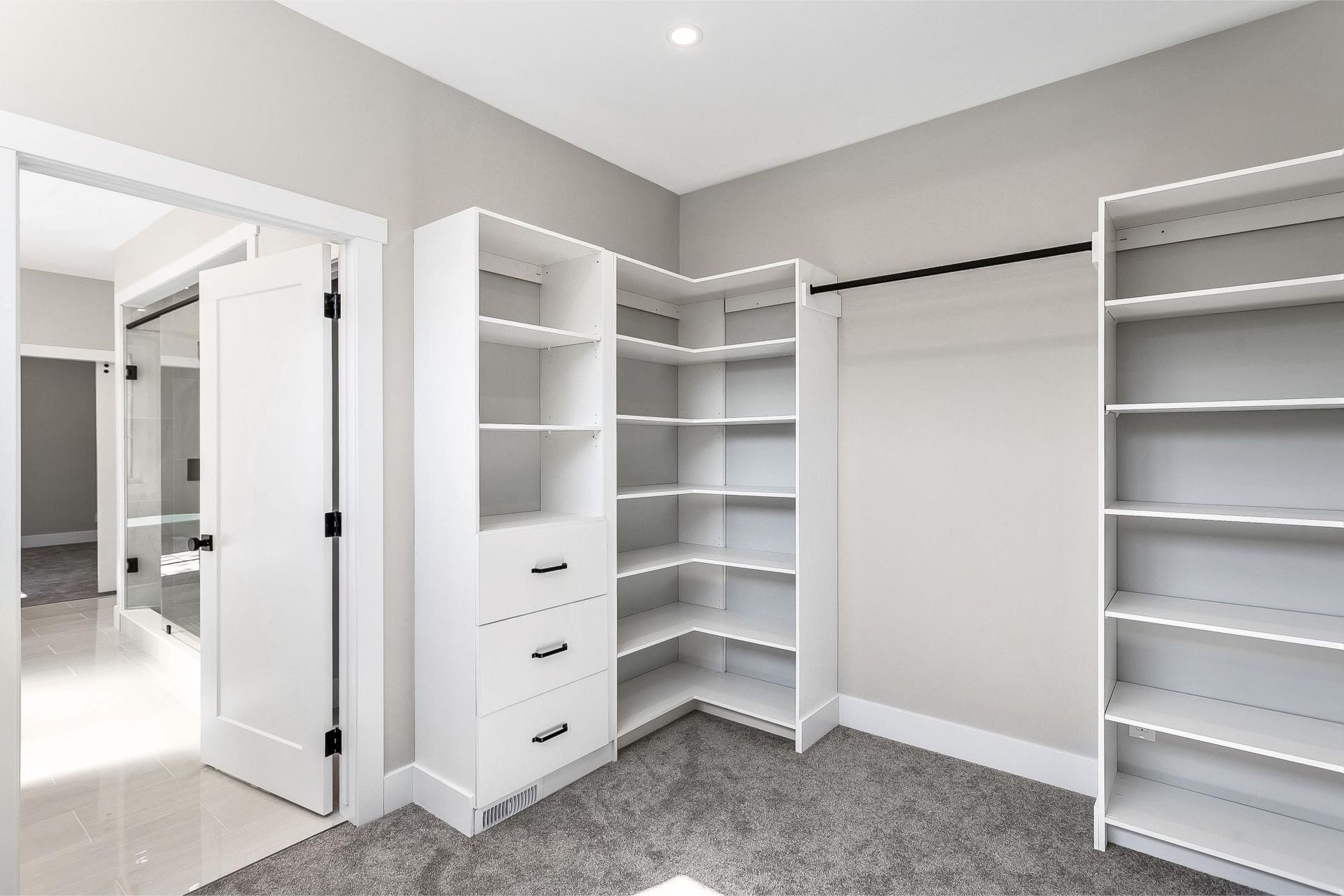 A walk-in closet featuring white built-in shelving, drawers, and a hanging rod against neutral walls with grey carpet.