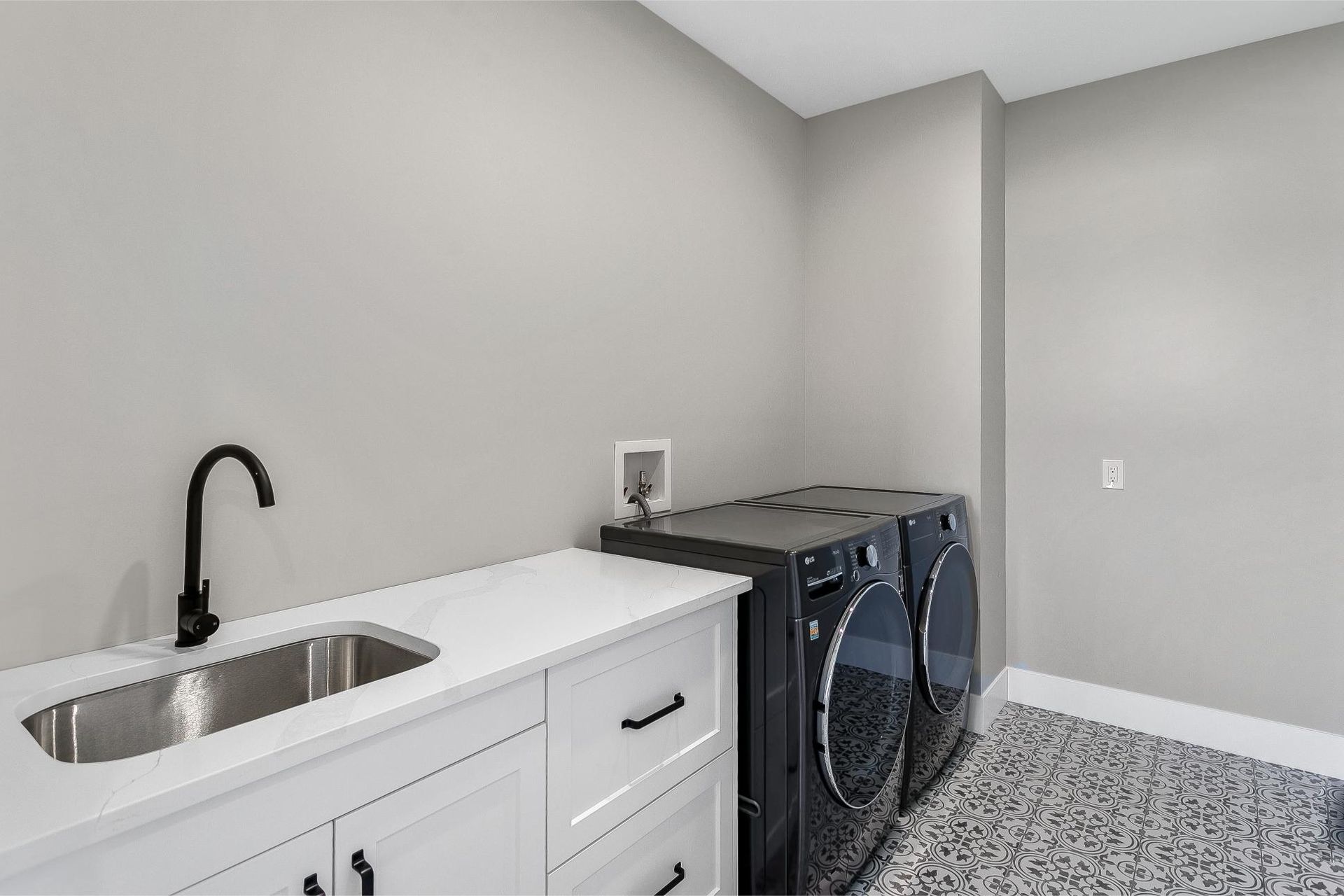 A laundry room with a white counter, black sink faucet, two black appliances, and gray patterned flooring.