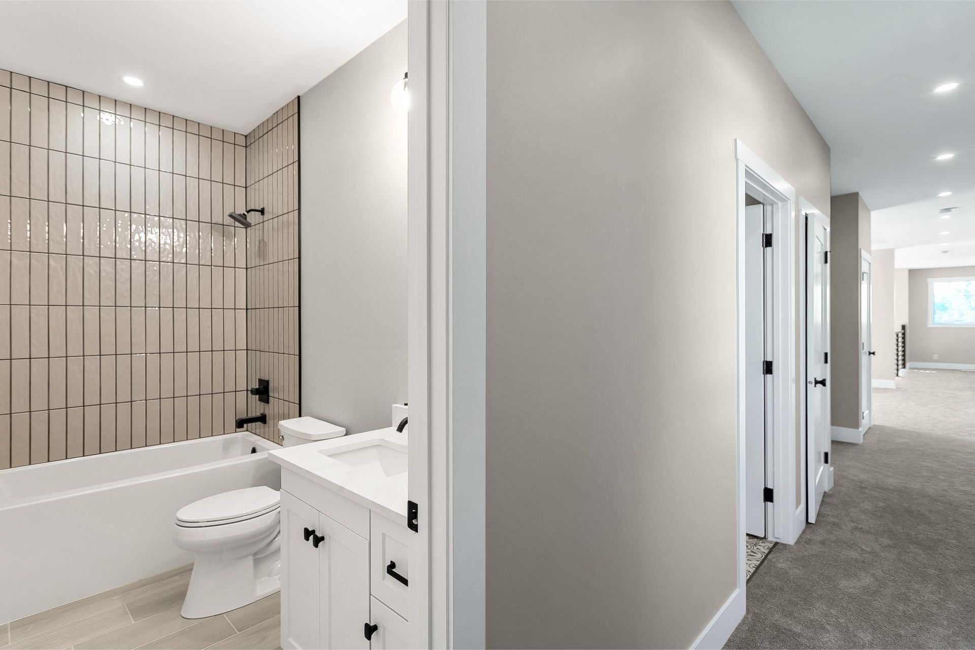 A modern bathroom with a white vanity and tiled shower next to a hallway with carpet and multiple white doors.