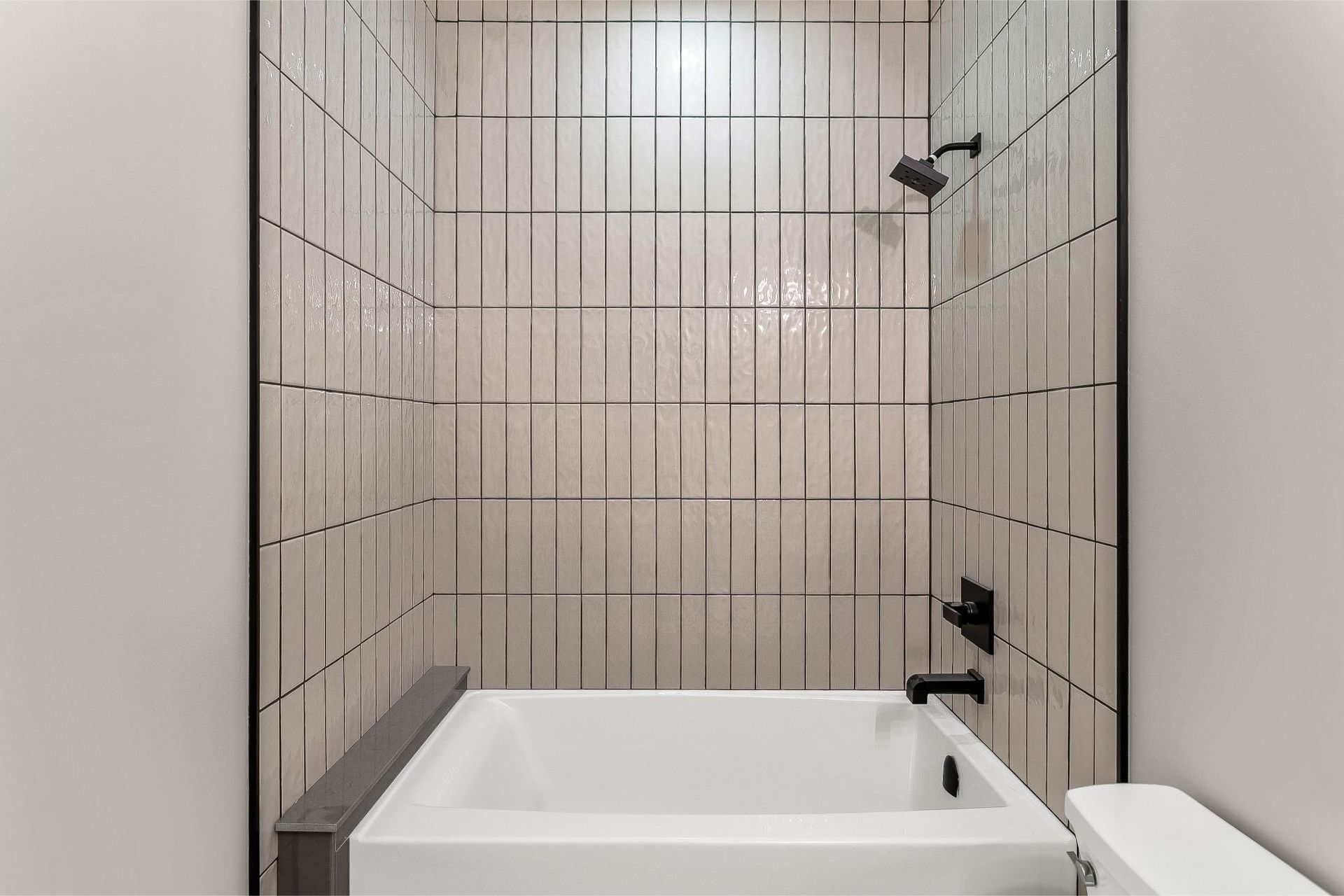 A modern bathtub and shower area with vertical, off-white rectangular tiles and black fixtures against a white wall.