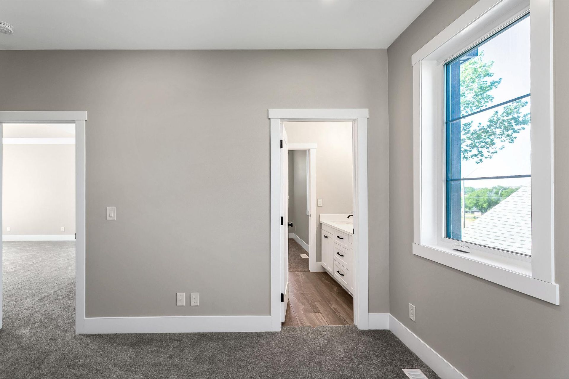 A neutral-toned room with gray carpet, white trim, a large window, and two doorways leading to interior spaces.