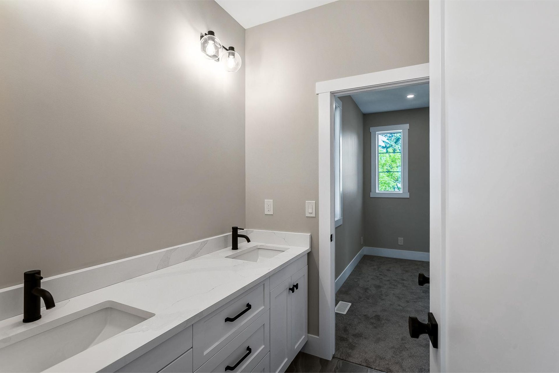 A modern bathroom vanity with dual sinks, white countertops, black fixtures, and light gray walls, leading to a bedroom.