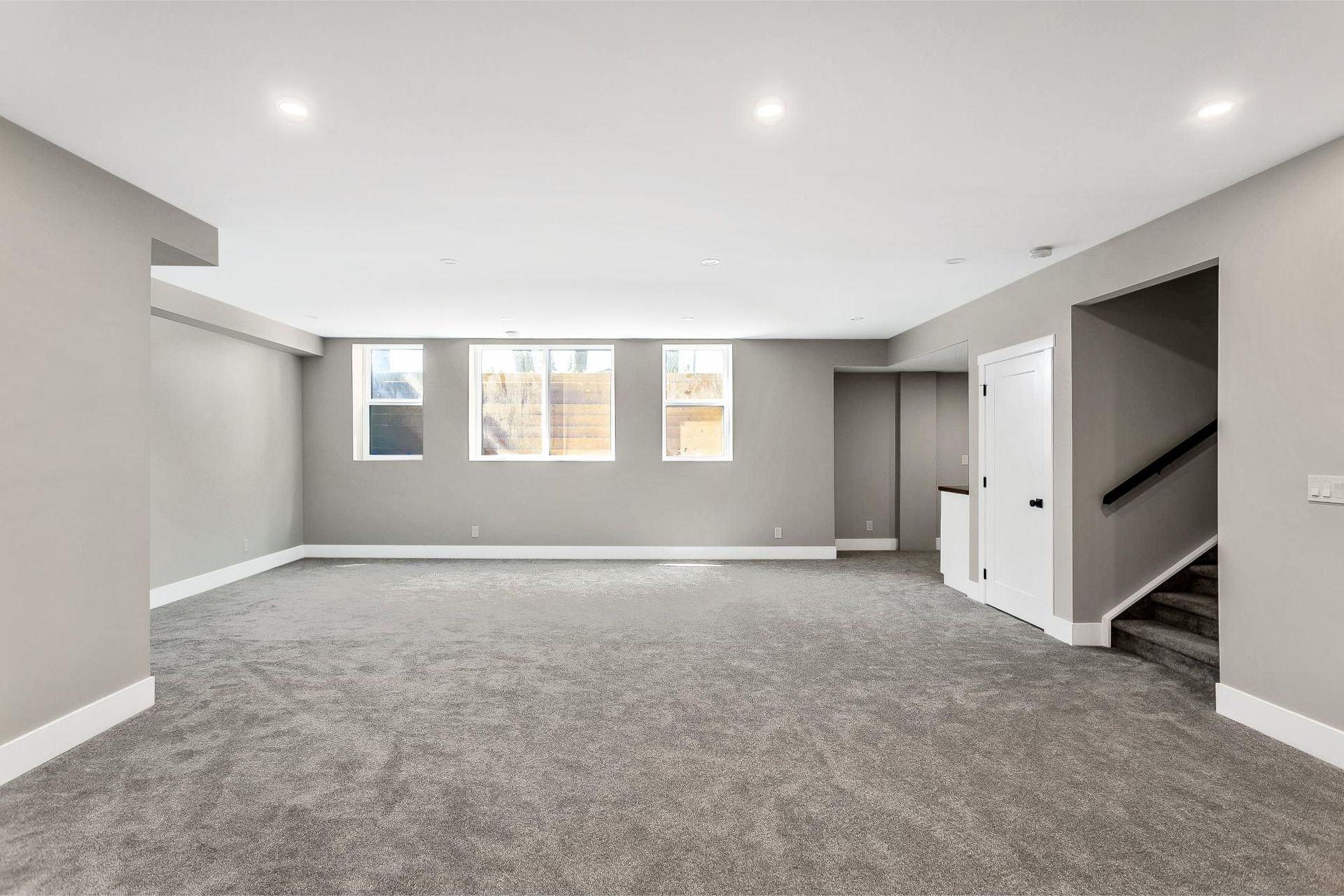 An empty basement room with grey carpet, light grey walls, three windows, and a staircase leading to an upper floor.