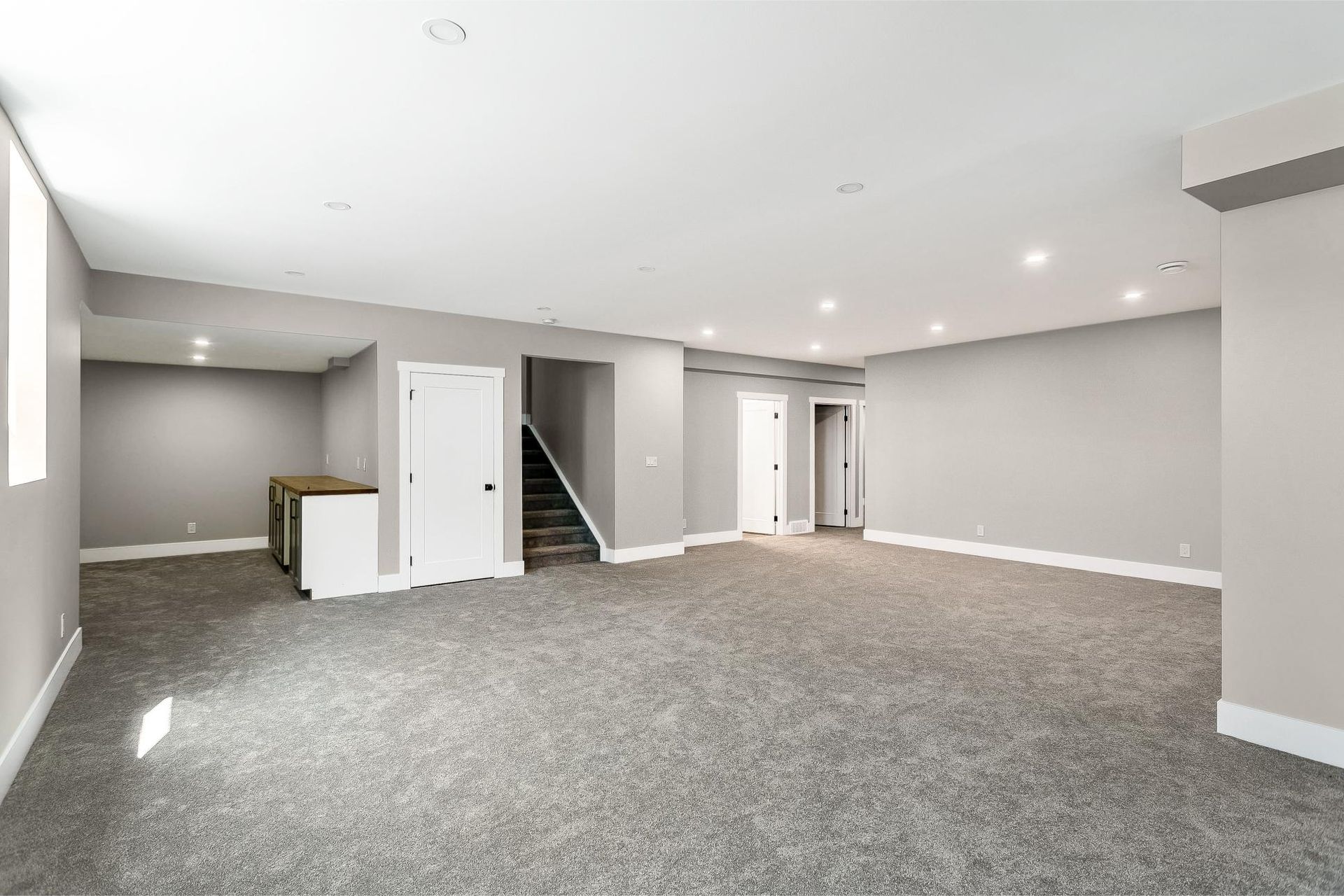 An open basement room with neutral walls, grey carpeting, recessed lighting, and a staircase leading upstairs.