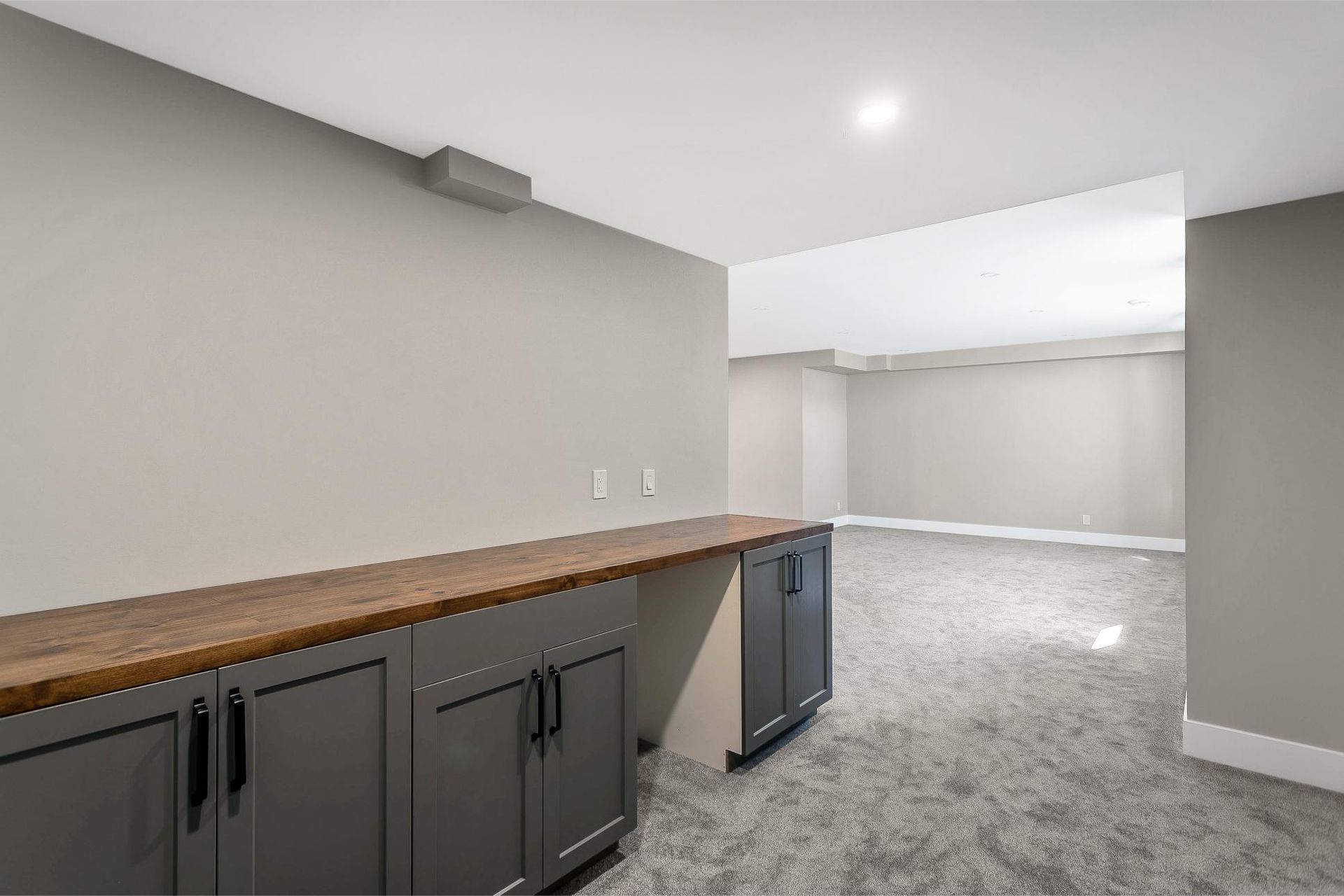 A basement interior featuring dark gray cabinets with a wooden countertop and light gray walls over gray carpet flooring.