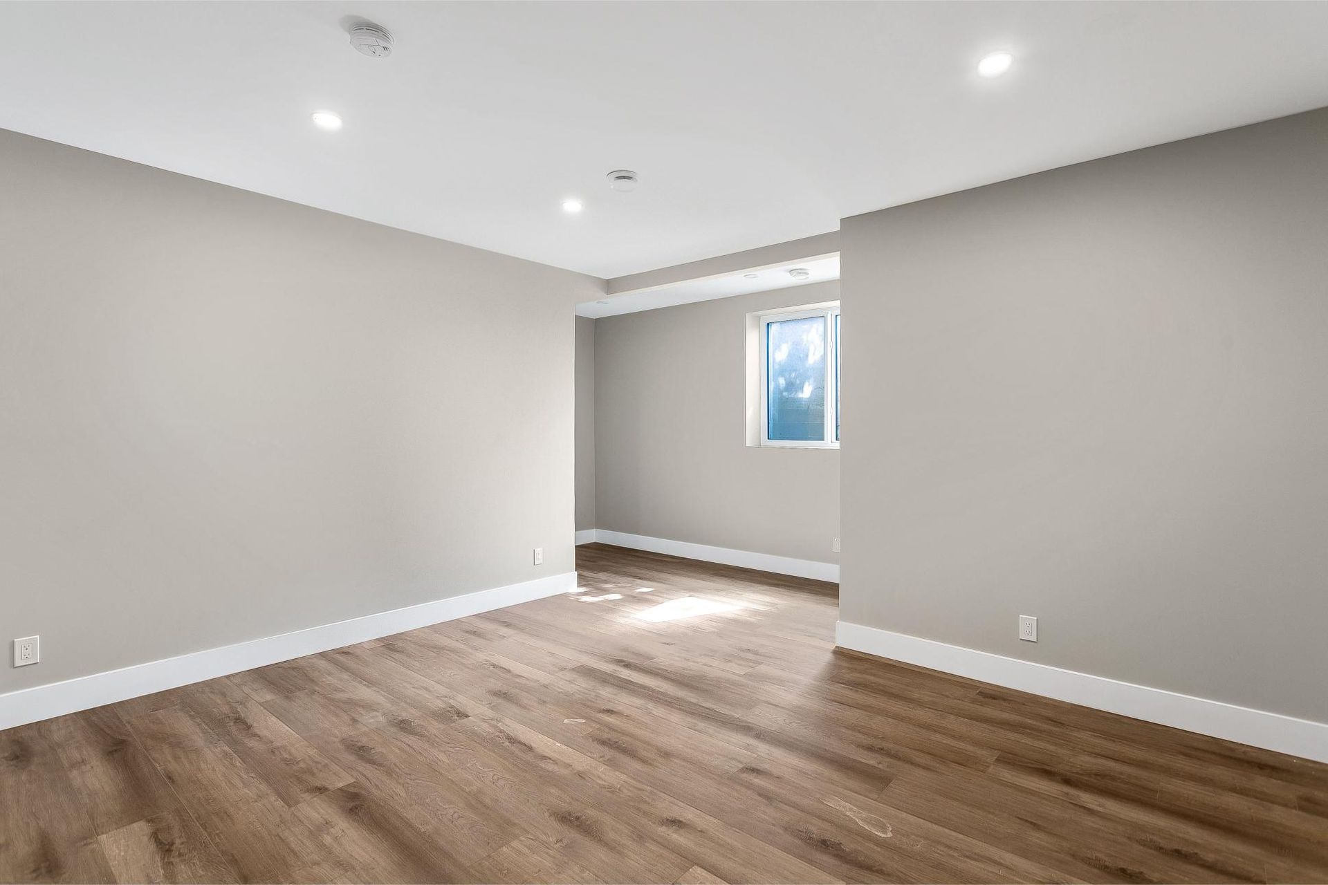 A vacant room with light grey walls, white trim, wood-look flooring, and a single window in the back corner.