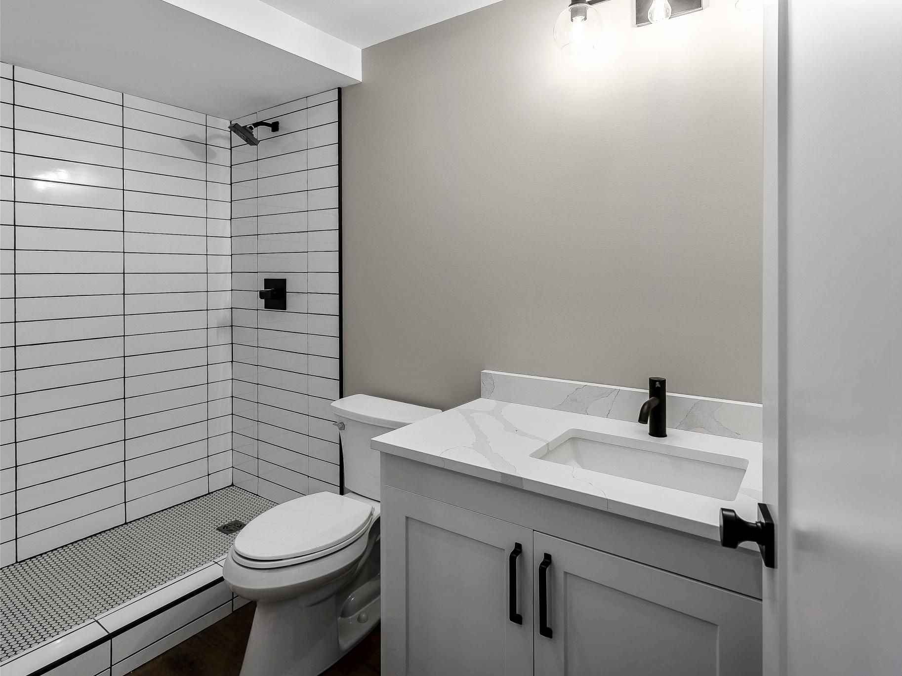 A modern bathroom with a white vanity, a toilet, and a walk-in shower featuring white tiled walls with metallic accents.