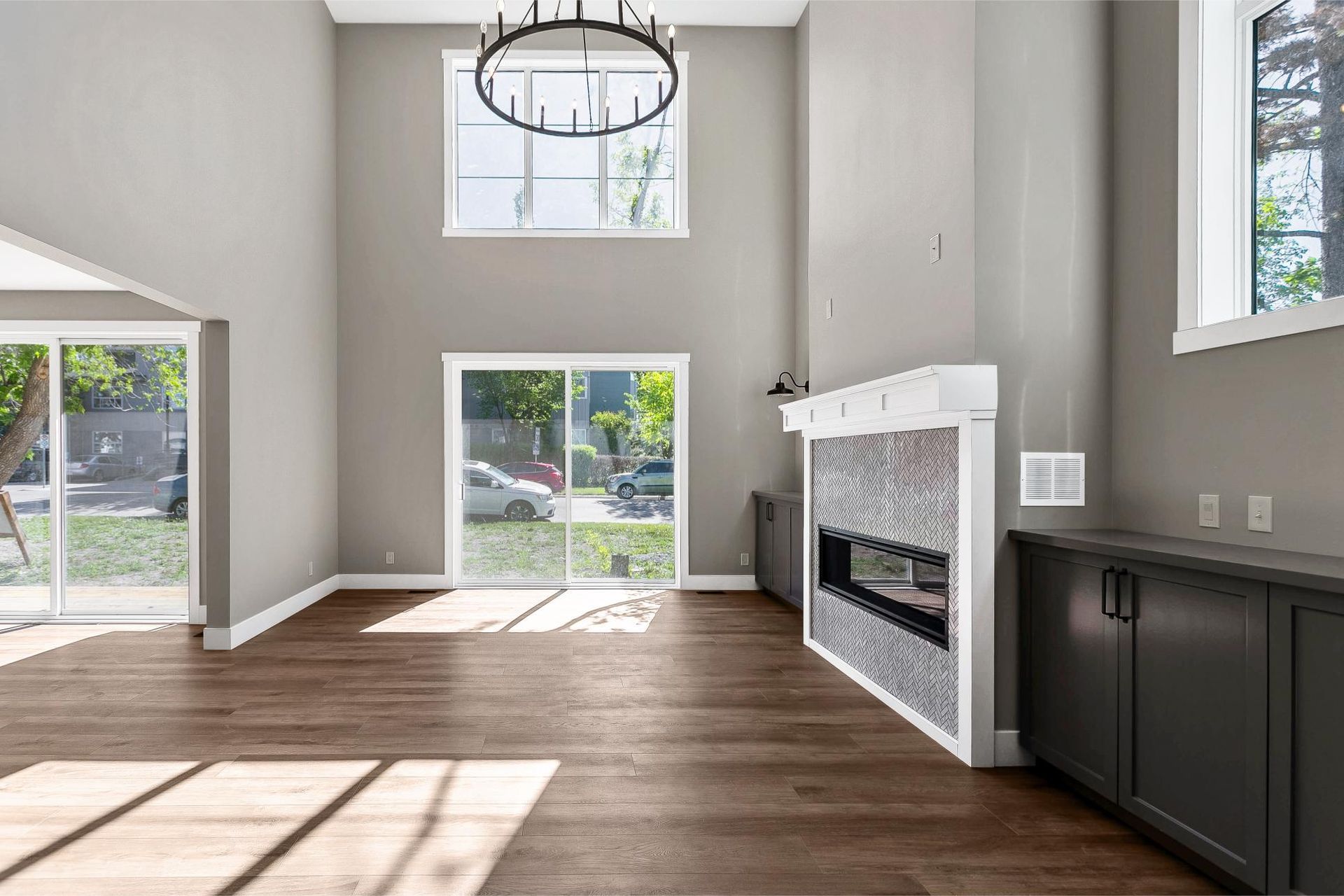 A modern, high-ceilinged living room with wood floors, a tiled fireplace, dark cabinets, and large windows.