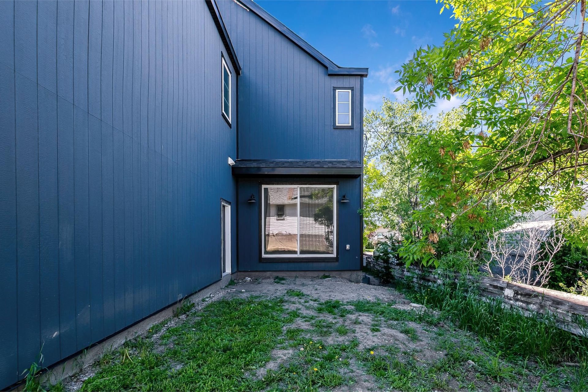 A side view of a modern, two-story house with dark blue vertical siding, a single window, and a green yard with trees.