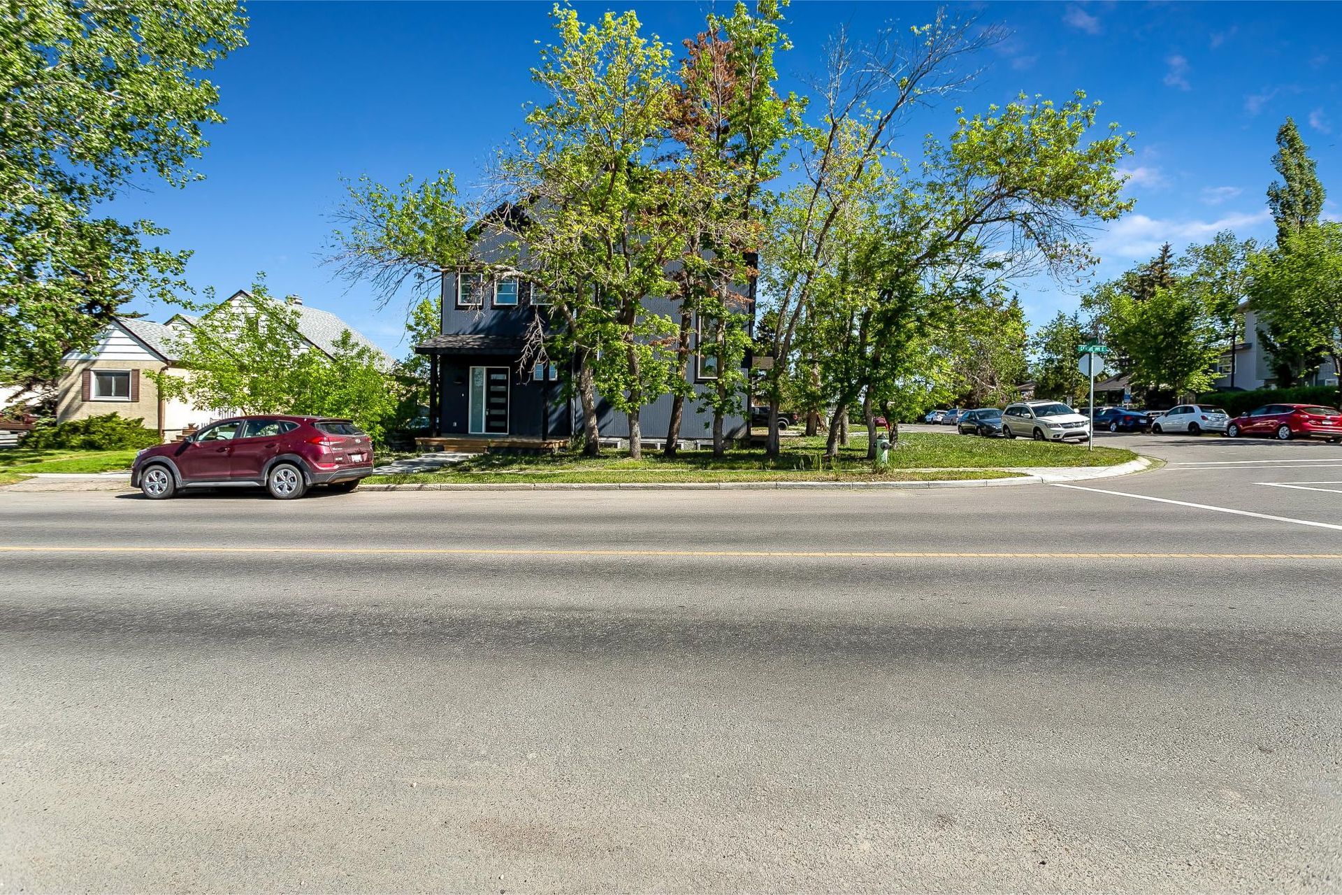 A dark-colored house set behind trees on a sunny day, with a red car parked on the street in the foreground.