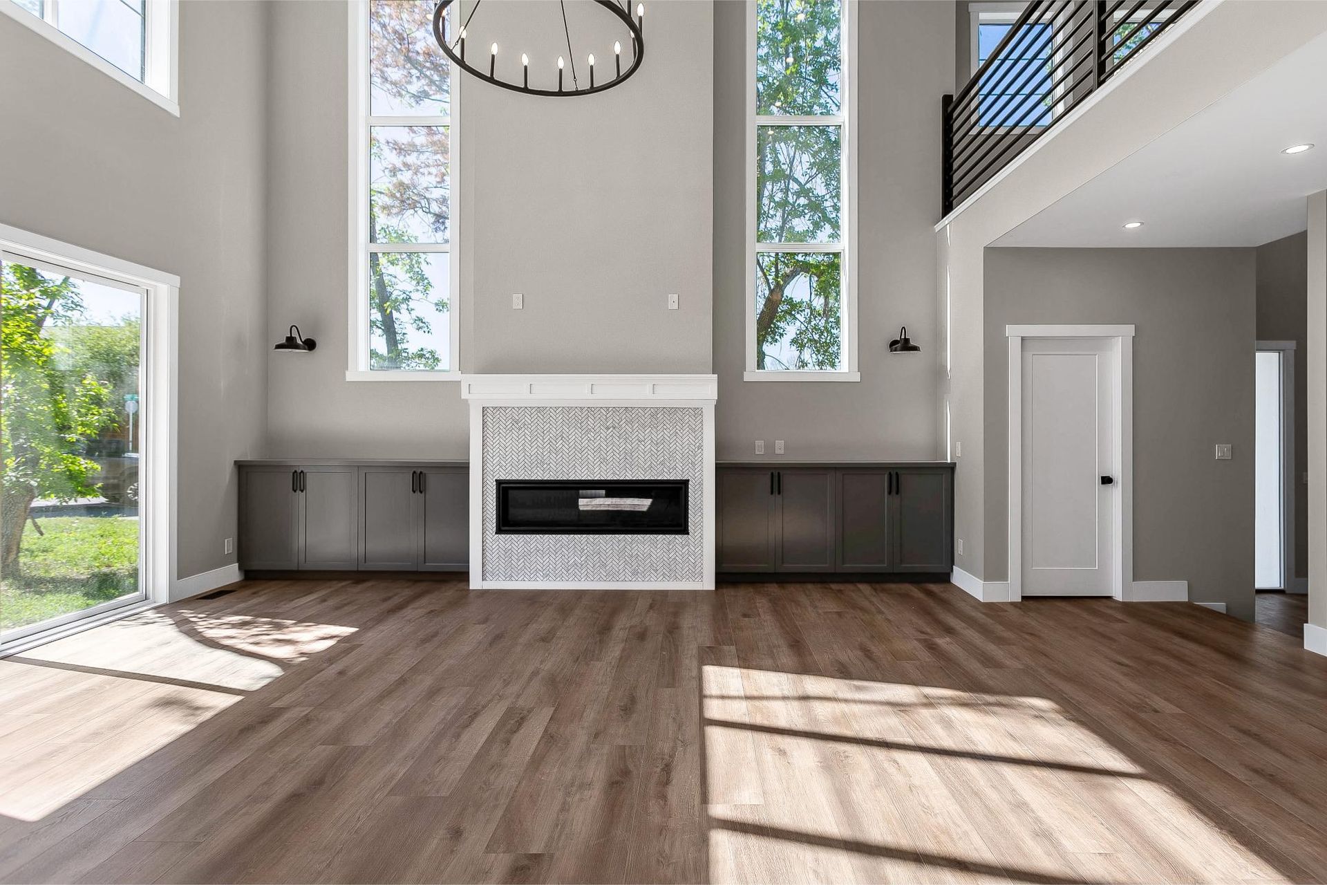 A modern, sunlit living room with high ceilings, large windows, a chevron-tiled fireplace, and dark built-in cabinetry.