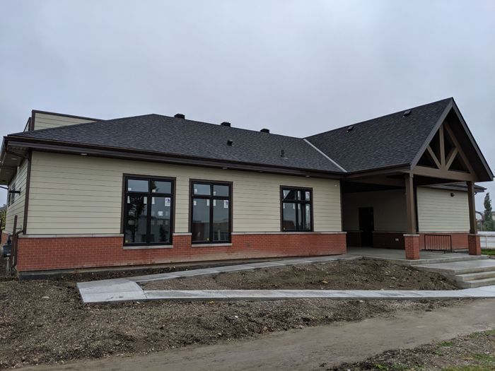 Newly built single-story building with beige siding, brick base, dark roof, and a covered front entryway under construction.