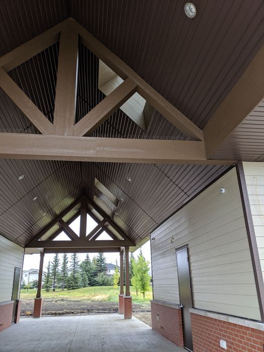 An outdoor covered walkway with dark brown beams, a sloped paneled ceiling, light siding, and a red brick base.