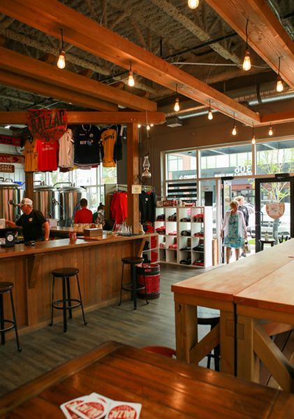 A brightly lit brewery taproom with wooden tables, bar seating, industrial metal tanks, and colorful shirts hanging.