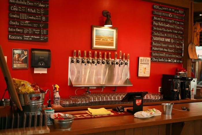 A bar counter with a row of beer taps on a bright red wall, featuring chalkboards listing drink menus on both sides.