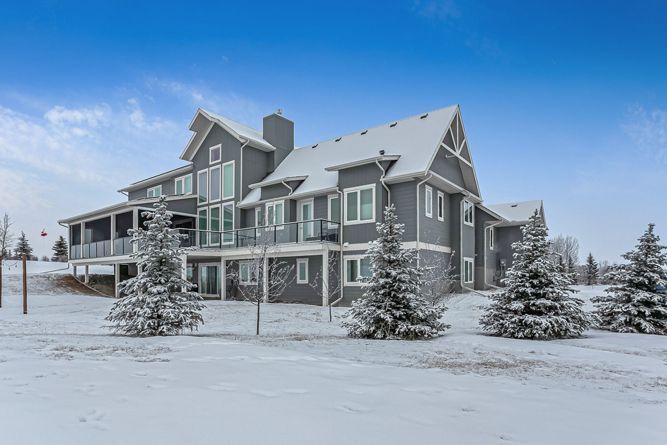 A large, multi-story gray house with a snowy roof, surrounded by snow-covered ground and evergreen trees under a blue sky.