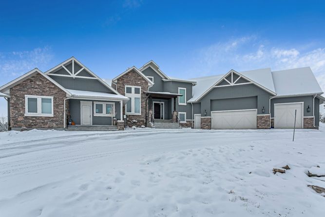 A large, two-story house with dark gray siding and stone accents, set in a snowy landscape under a clear blue sky.