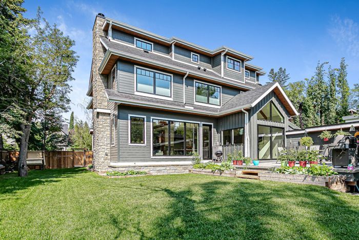 A grey, multi-story house with a stone chimney and large windows, set against a green lawn under a clear blue sky.