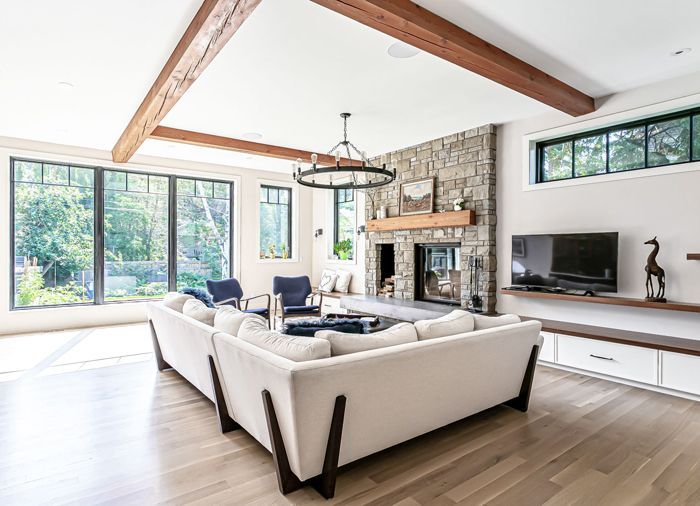 A modern living room with a white sectional sofa, stone fireplace, wooden ceiling beams, and large windows.