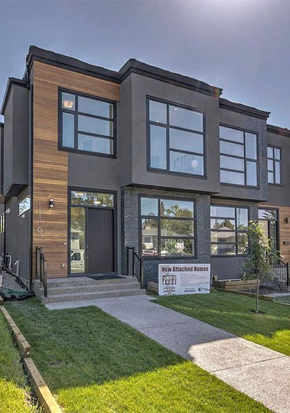 A modern two-story townhouse with dark grey stucco, wood siding accents, large rectangular windows, and a paved walkway.