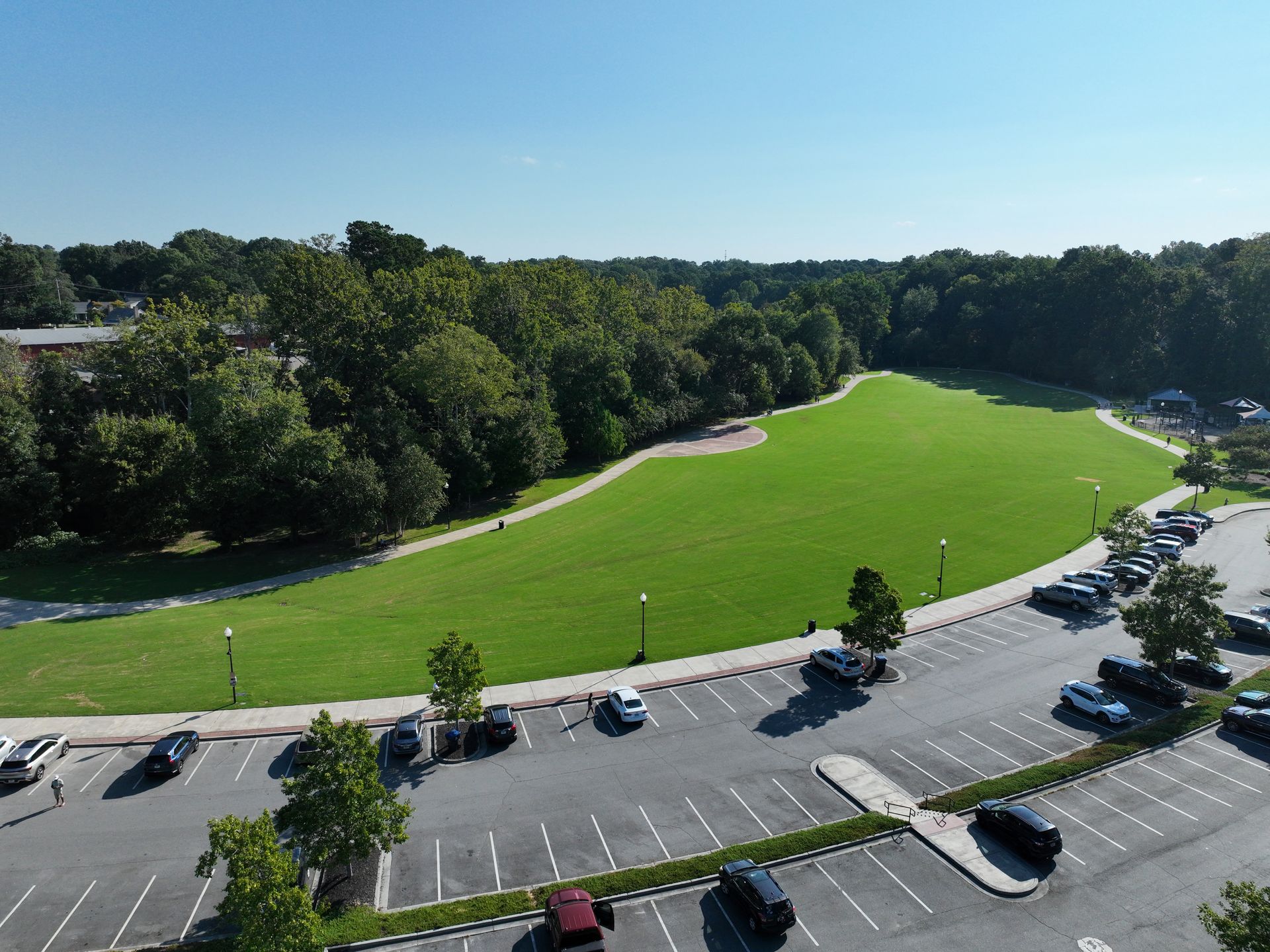 Aerial view of a large green park beside a parking lot, surrounded by trees under a clear sky