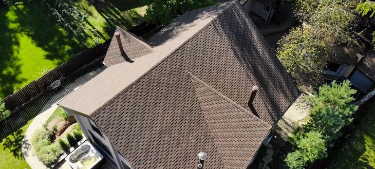 Aerial view of a brown shingled house roof surrounded by trees and a yard