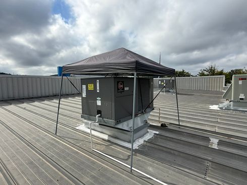 Rooftop HVAC unit under a metal canopy on a corrugated roof, with cloudy sky overhead.