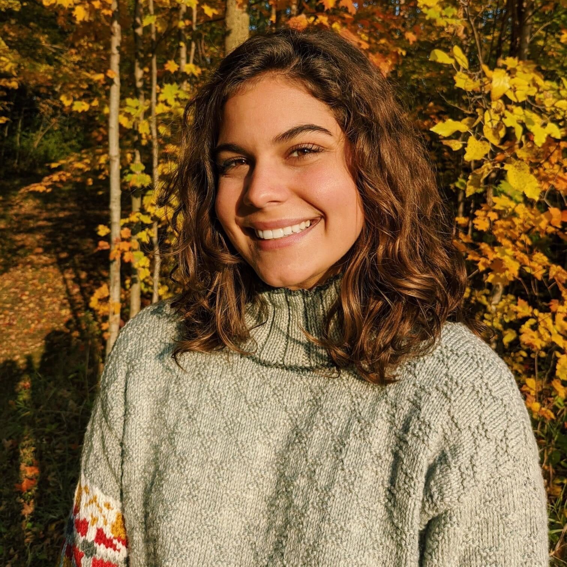 Person smiling outdoors, wearing a gray sweater, standing in front of fall foliage.