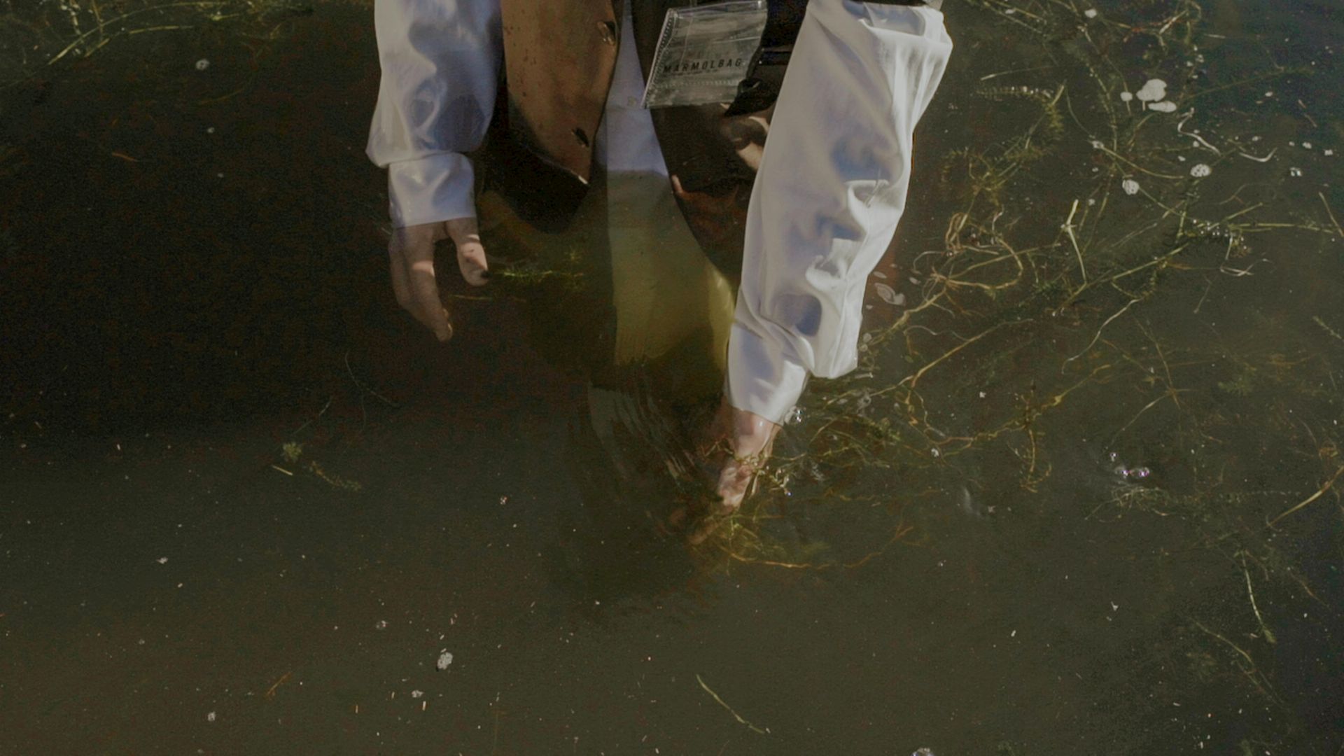 Person steps barefoot into murky water, wearing white shirt and pants.