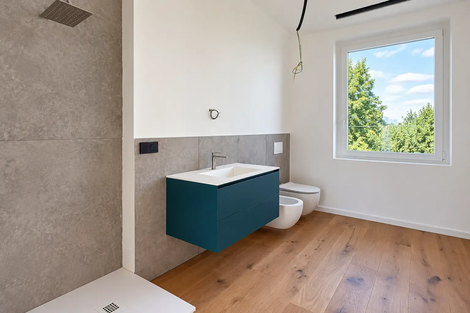 Bagno moderno con mobile lavabo color verde petrolio, WC bianco, doccia a filo pavimento e un’ampia finestra con vista sugli alberi
