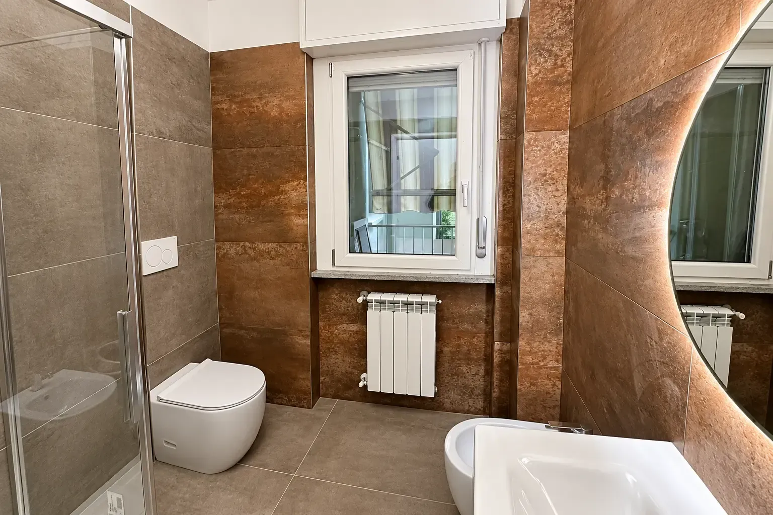 Bagno moderno con grandi pareti rivestite di piastrelle marroni, WC sospeso, finestra, termosifone e bordo della vasca bianco