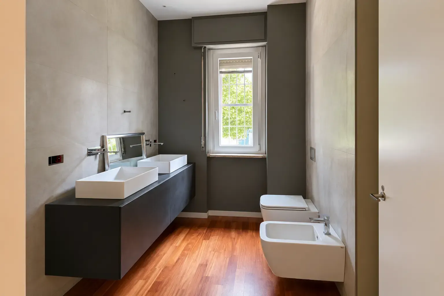 Bagno moderno con doppio lavabo, mobile scuro, WC bianchi e una finestra sopra il pavimento in parquet