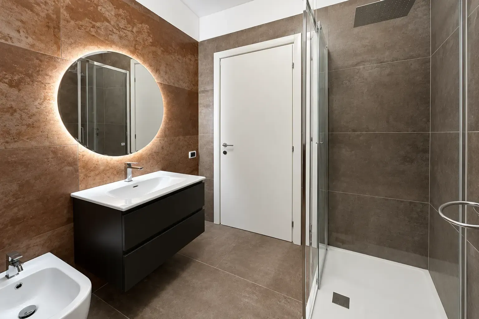 Bagno moderno con piastrelle marroni, specchio rotondo, mobile lavabo nero, WC bianco e box doccia in vetro