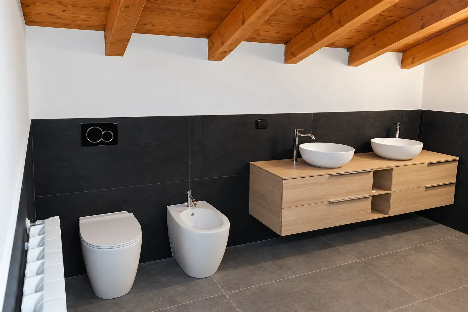 Bagno moderno con piastrelle nere, due lavabi a parete, bidet e un mobile lavabo in legno sotto travi a vista