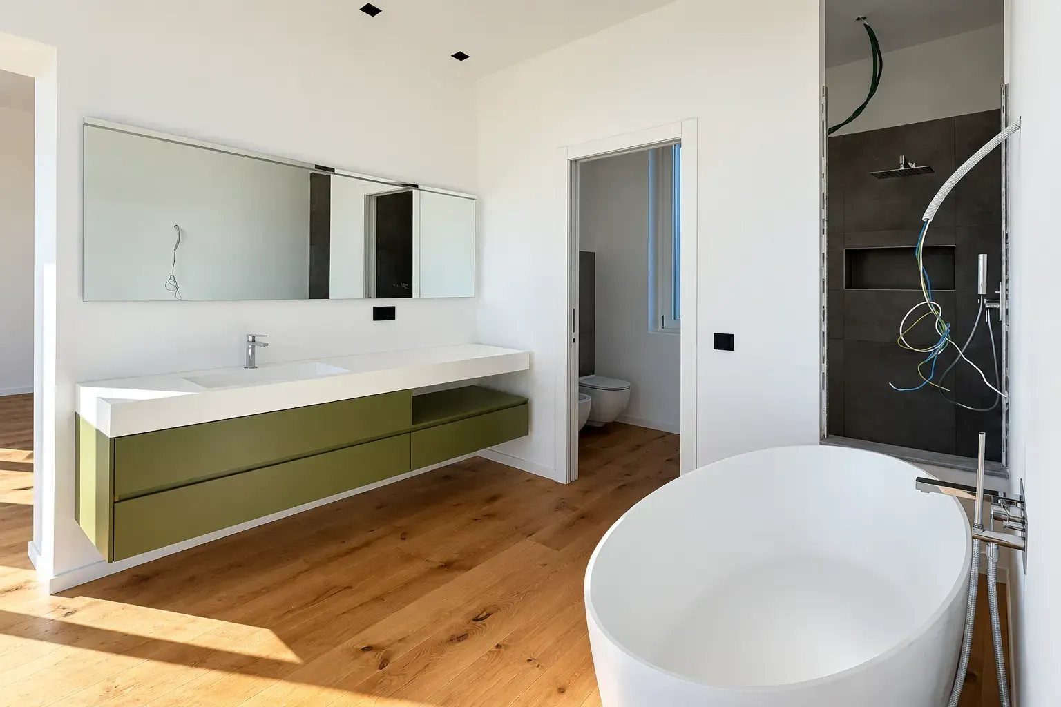 Bagno moderno con mobile lavabo verde, grande specchio, pavimento in legno e vasca bianca freestanding