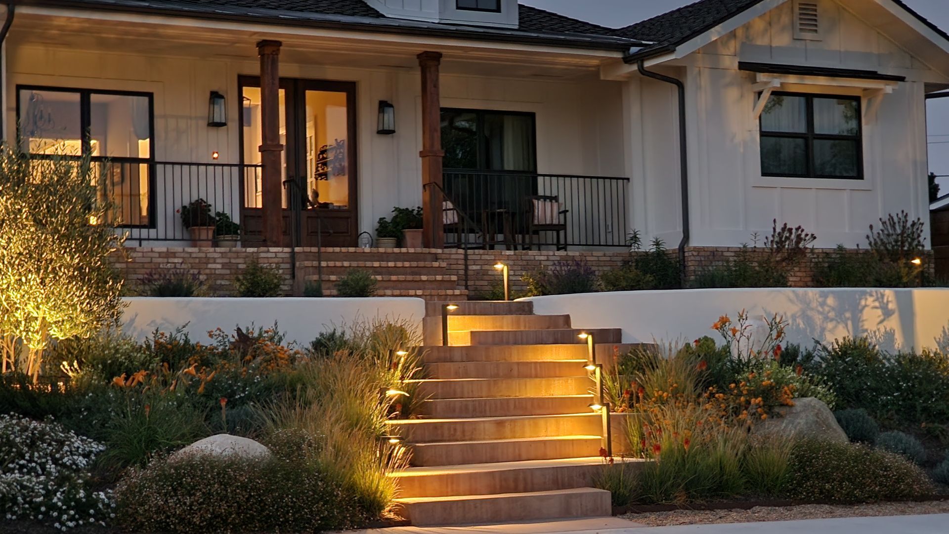 Front porch of a house at dusk with lit steps, warm exterior lights, and landscaped garden beds.