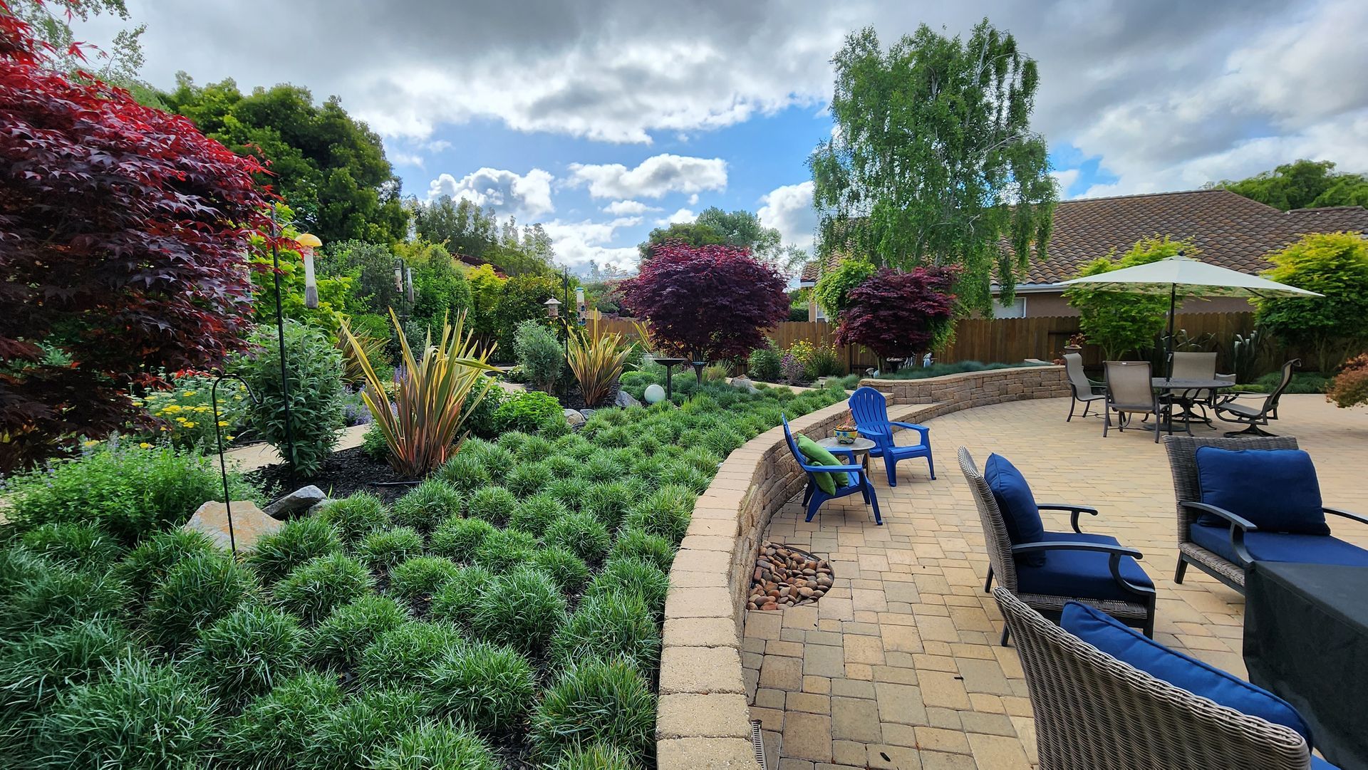 Lush garden patio with blue lounge chairs, paved path, and colorful flowers under a partly cloudy sky