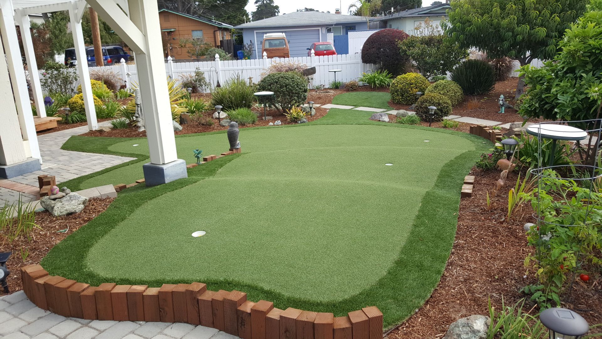 Backyard putting green with artificial turf and brick edging, bordered by garden plants and a patio
