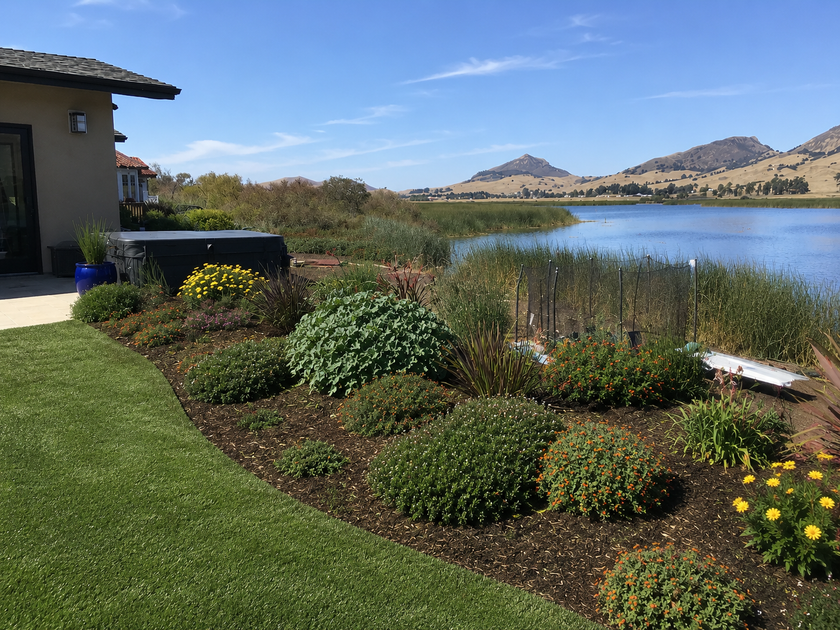 Lakefront garden with colorful flower beds beside a house, water, and distant hills under a clear sky