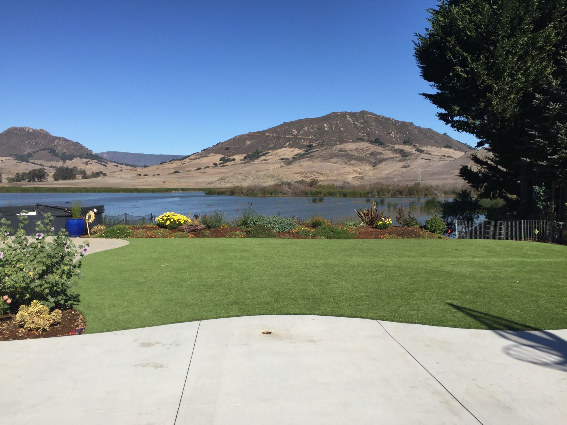 Backyard lawn and patio overlooking a lake and tan hills under a clear blue sky.