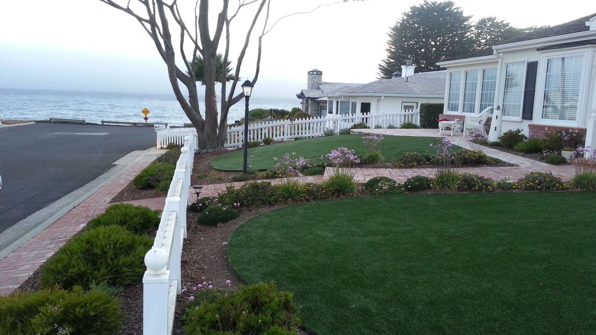 White coastal house with manicured lawn and garden beside a road overlooking the ocean