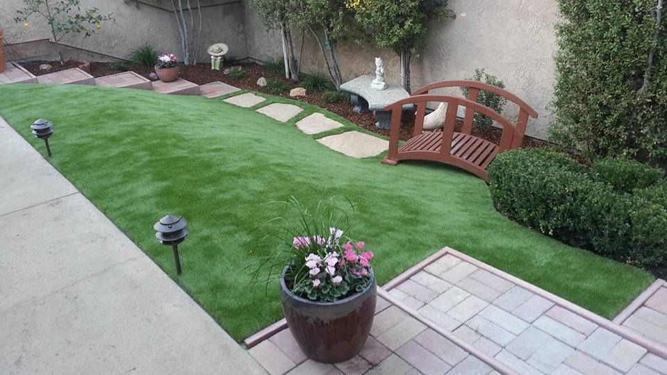 Backyard patio with green lawn, stepping stones, small wooden bridge, and flower pots
