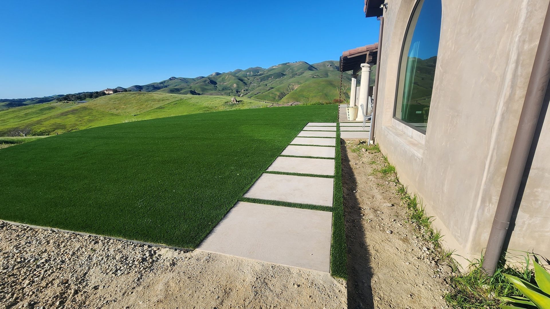 Stone walkway beside a beige house, bordered by bright green lawn and hills under a clear blue sky